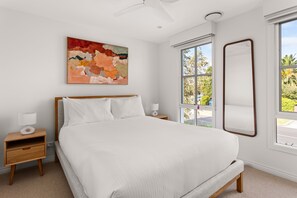 A bedroom with a bed, a nightstand, a lamp, a ceiling fan, a window with a view, and a painting on the wall.