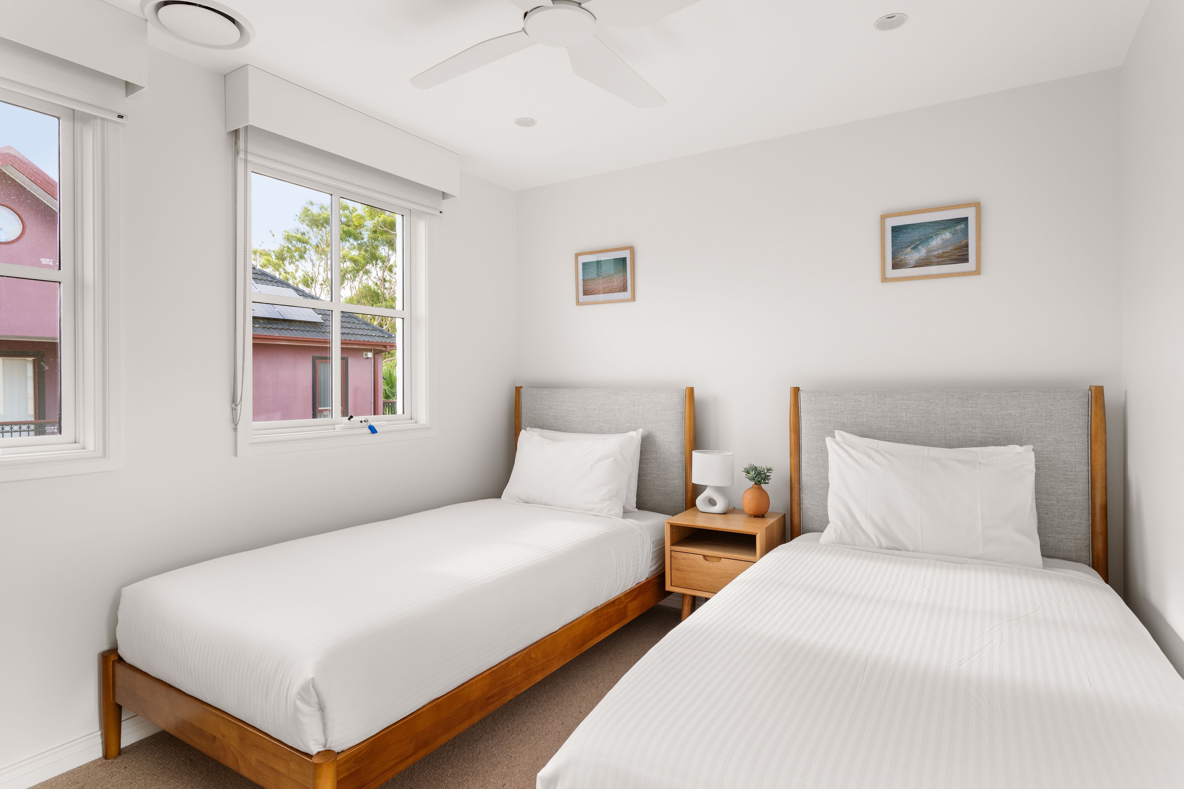 A hotel room with two single beds, a nightstand, a ceiling fan, and two framed pictures on the wall.