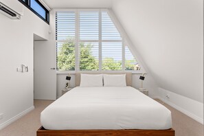 Un dormitorio con techo inclinado, un ventanal con persianas, una cama con ropa de cama blanca, dos mesitas de noche con lámparas y una pequeña mesita auxiliar con una planta.