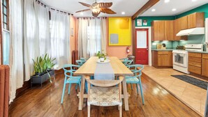 Un coin repas avec une table en bois et des chaises bleues, un ventilateur de plafond, et une cuisine dotée de meubles en bois et d’une cuisinière.