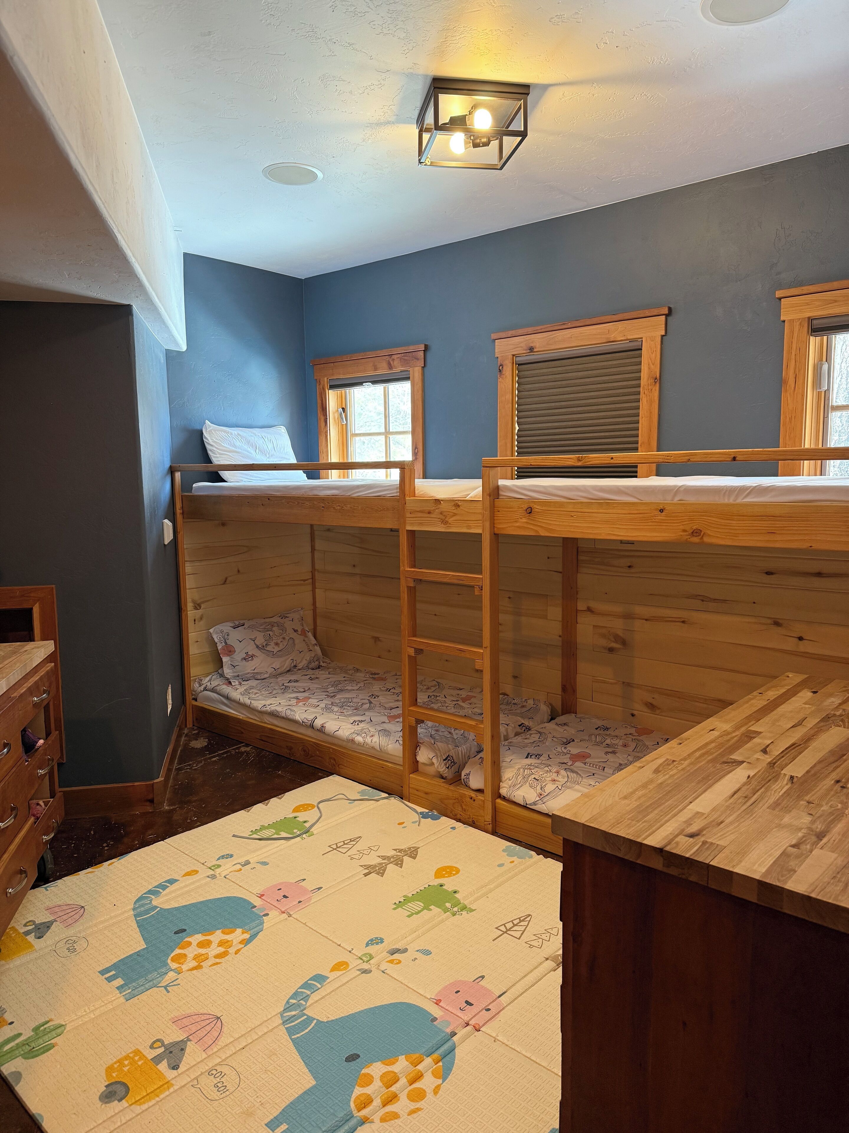 A bunk bed room with a wooden ladder, a small wooden dresser, and a window with blinds.