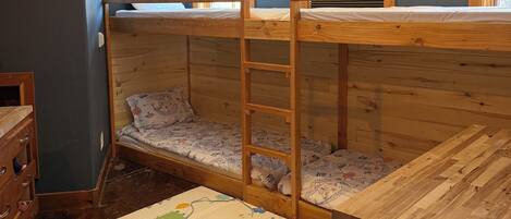 A bunk bed room with a wooden ladder, a small wooden dresser, and a window with blinds.