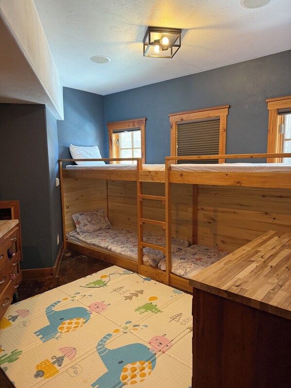 A bunk bed room with a wooden ladder, a small wooden dresser, and a window with blinds.