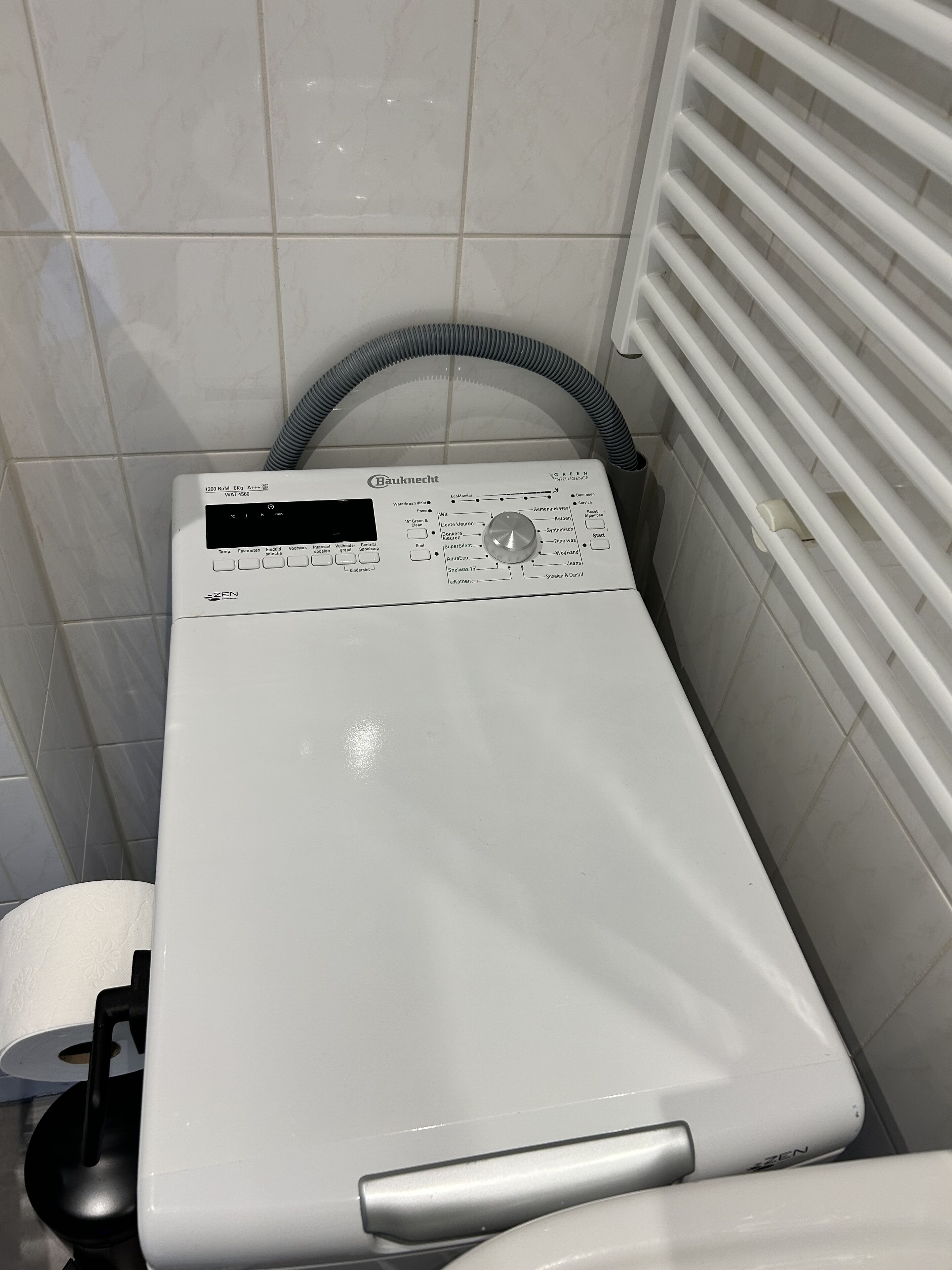 A white Bauknecht washing machine in a bathroom with white tiles and a window with blinds.