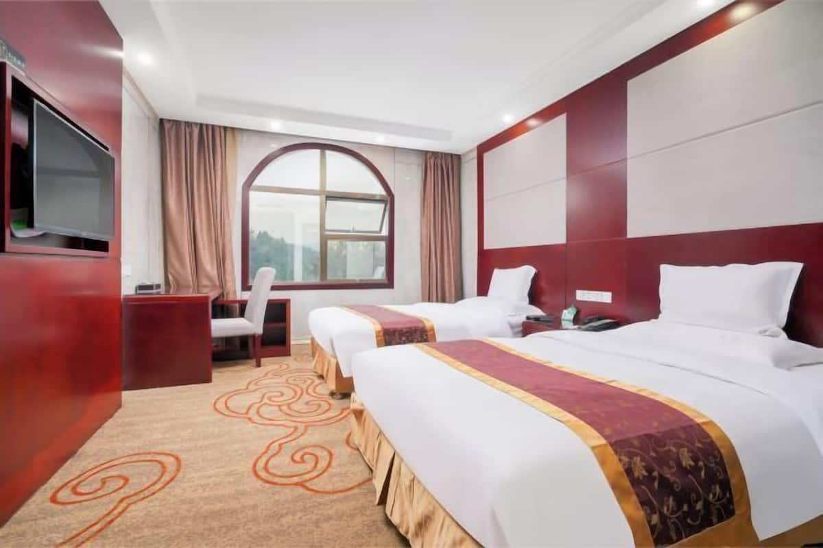A hotel room with two beds, a TV, a desk, and a large window.
