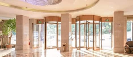 Un vestíbulo de hotel con piso de mármol pulido, una lámpara de araña grande y puertas de vidrio que dan al exterior.