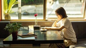Une personne est assise à une table près d’une fenêtre, avec une plante en pot et un vase contenant une fleur.
