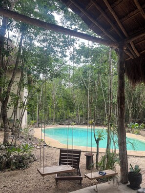 A wooden swing chair overlooking a turquoise pool, surrounded by lush greenery and a thatched roof structure. - Sak lool (Francisco Uh May)