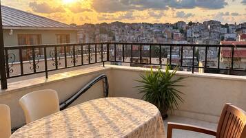 Un balcon avec une table et des chaises, d’où l’on peut admirer le panorama urbain au coucher du soleil.