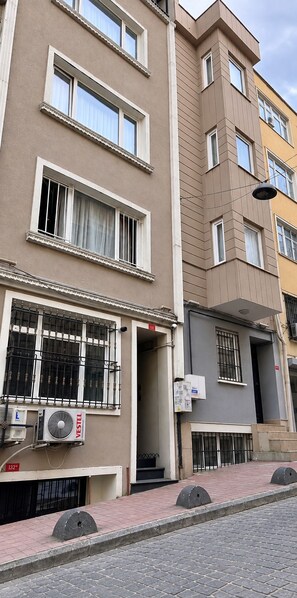 A multi-story building with balconies, a visible air conditioning unit, and a street with bollards.