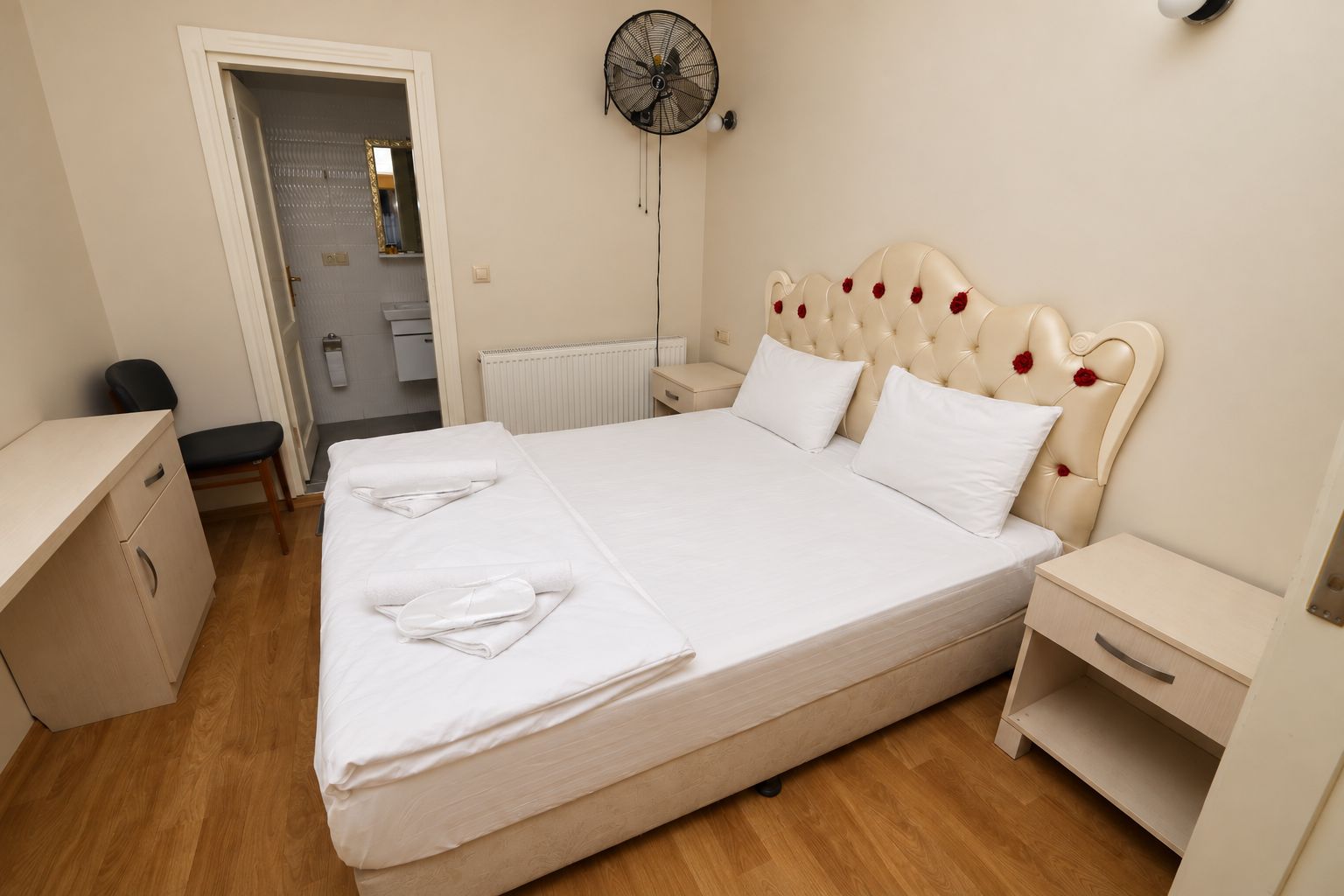 Un lit bien fait, avec du linge de lit blanc, une table de chevet, une commode, un ventilateur fixé au mur et une porte donnant sur une salle de bain.