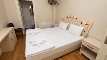 Un lit bien fait, avec du linge de lit blanc, une table de chevet, une commode, un ventilateur fixé au mur et une porte donnant sur une salle de bain.
