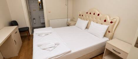 Una cama bien hecha con sábanas blancas, una mesita de noche, un ventilador fijado a la pared y una puerta que da al baño.