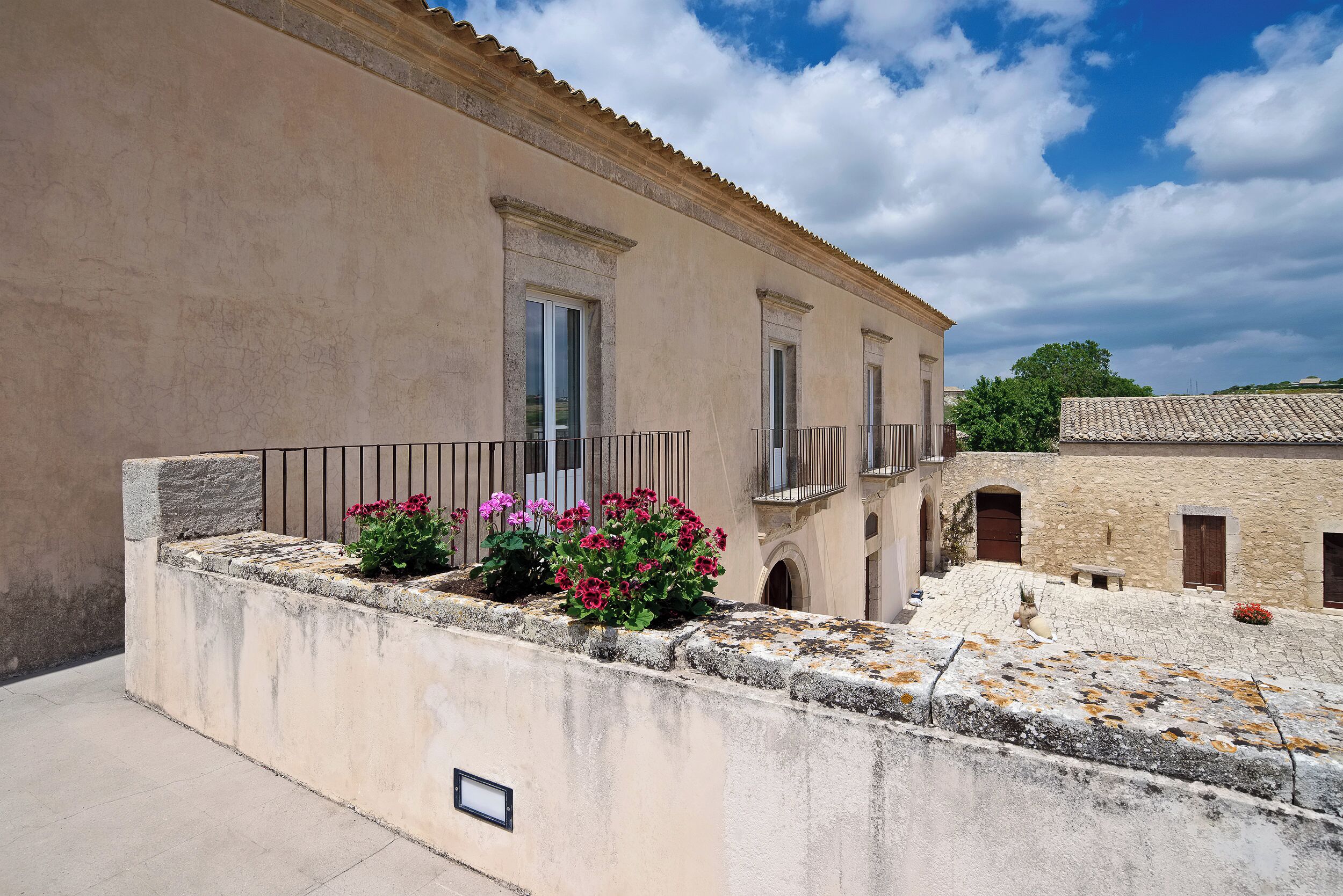A building with a stone facade, a balcony with metal railings, and a flower bed with pink flowers.
