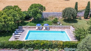 A rectangular swimming pool with a stone border, surrounded by lounge chairs and umbrellas, set against a backdrop of a field and trees. - Scifazzo, ancient Sicilian villa with pool (Ragusa)