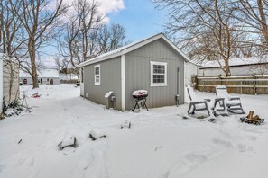 Une cour arrière enneigée avec un abri de jardin gris, un barbecue et une terrasse recouverte de neige.