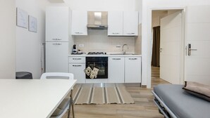 A compact kitchen with white cabinets, a stove, and a sink. There is a dining table with a striped rug, and a bed is visible in the adjacent room.