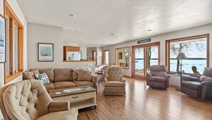 A spacious living room with a large sofa, armchairs, and a coffee table. There are multiple windows offering views of a lake. - Relaxing Lakefront Cottage, Kayaks, Bikes, Fishing (Sturgeon Bay)