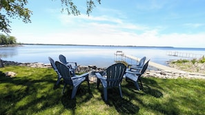 A lakeside seating area with Adirondack chairs and a table, overlooking a calm lake and distant shoreline.