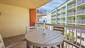 A balcony with a round table, two chairs, a bottle of wine, and two wine glasses. The balcony overlooks a building with multiple balconies and windows. - Oceanfront | Heated Pools + Hot Tub | King | W/D | Cable (Orange Beach)