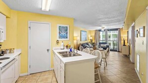 A compact kitchen with a central island, a dining area with a table and chairs, and a view of the outside through a glass door. - Oceanfront | Heated Pools + Hot Tub | King | W/D | Cable (Orange Beach)