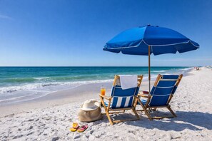 A beach scene with two blue beach chairs under a blue umbrella, a straw hat, and a pair of flip-flops on the sand.