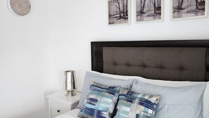 Una cama bien hecha con edredón blanco, dos almohadas decorativas, una lámpara de noche y una lámpara de pared. Tres cuadros enmarcados de un paisaje invernal están colgados sobre la cama.