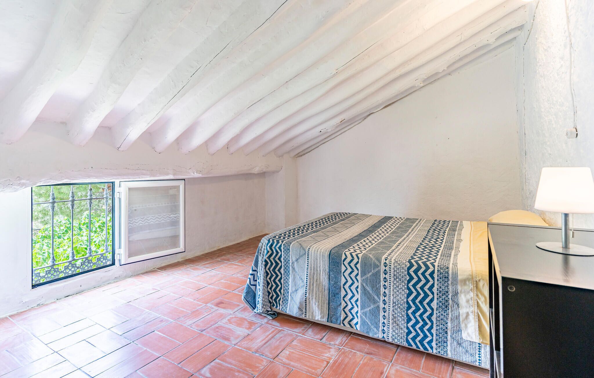 Ein kleines, helles Schlafzimmer im Dachgeschoss mit einem Bett, einem Fenster mit Blick auf Grünflächen und einer Lampe auf einem Tisch.