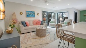 A modern living room with a white sofa, a dining table, wicker chairs, and a large window. - Sand & Sea Oasis: Condo with Private Beach Access (Holmes Beach)