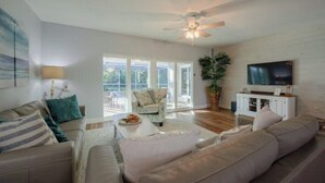 A spacious living room with a large sofa, a coffee table, and a television on a white cabinet. There is a ceiling fan and a large window providing natural light. - Coastal Oasis: Cheerful 3Bedroom, Pool, Large Yard (Bradenton)