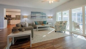 A modern living room with a sofa, coffee table, and armchair, a ceiling fan, and large windows offering a view of the outdoors. - Coastal Oasis: Cheerful 3Bedroom, Pool, Large Yard (Bradenton)