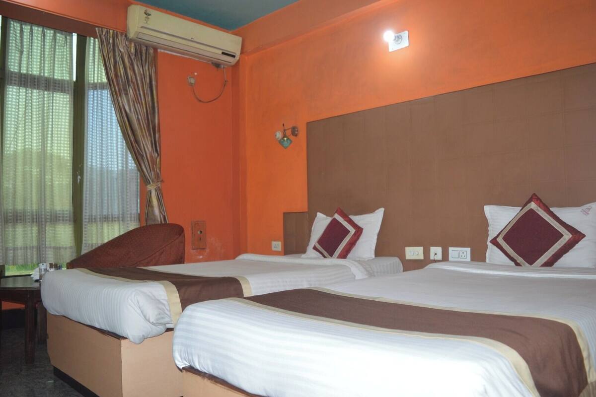 A hotel room with two beds, an air conditioning unit, a window with curtains, and a wall-mounted light fixture.