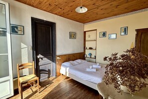 Quarto com teto de madeira, cama com roupa de cama branca, uma cadeira e um vaso com planta.