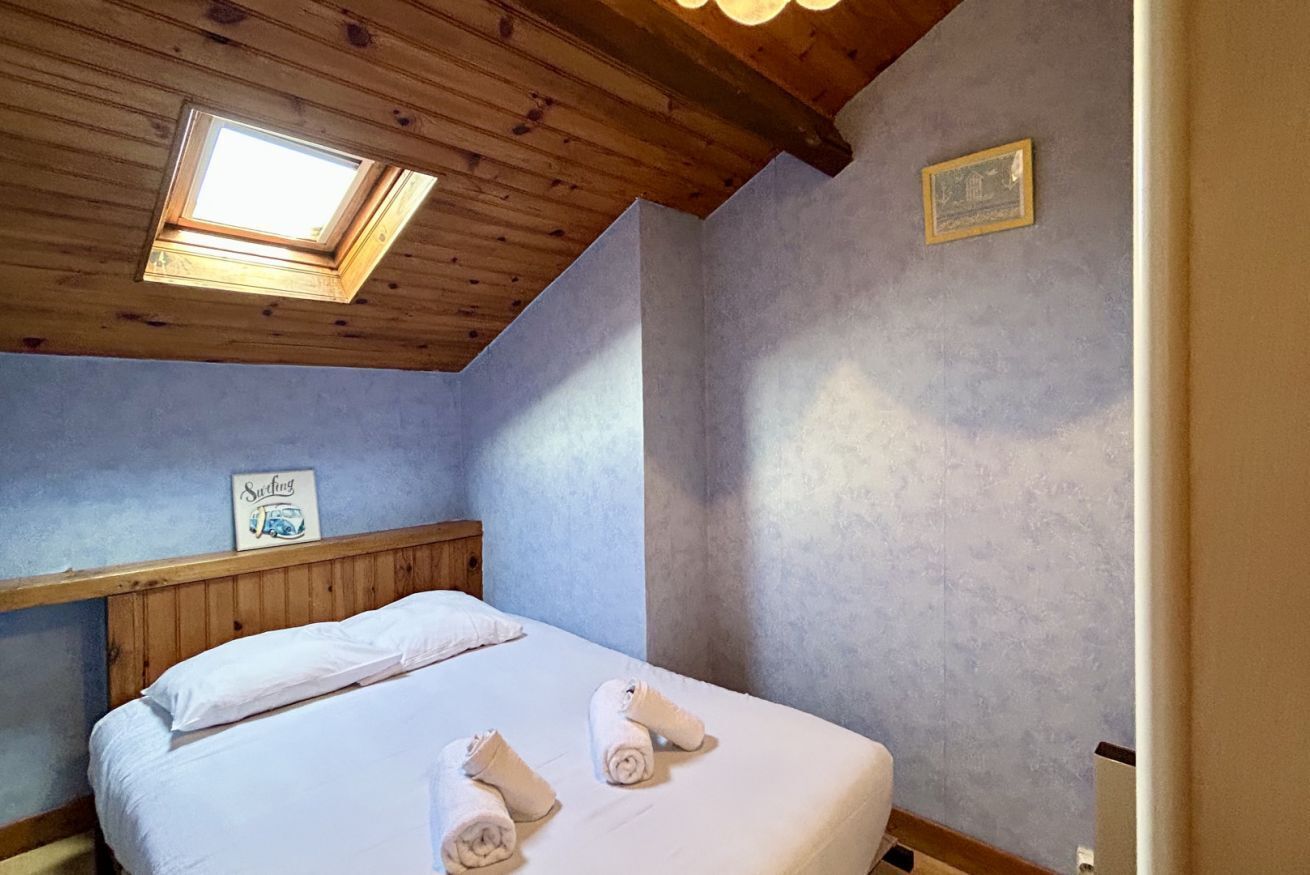Une chambre avec un plafond en bois, une lucarne, un lit avec du linge de maison blanc et un petit tableau encadré accroché au mur.