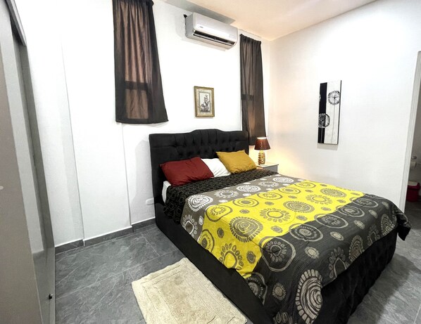 A bedroom with a bed, a nightstand, an air conditioning unit, and a door.