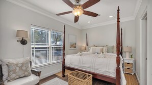 A bedroom with a four-poster bed, a ceiling fan, and a large window with blinds. - Charming Coastal Cottage on St. Simons Island (St. Simons Island)