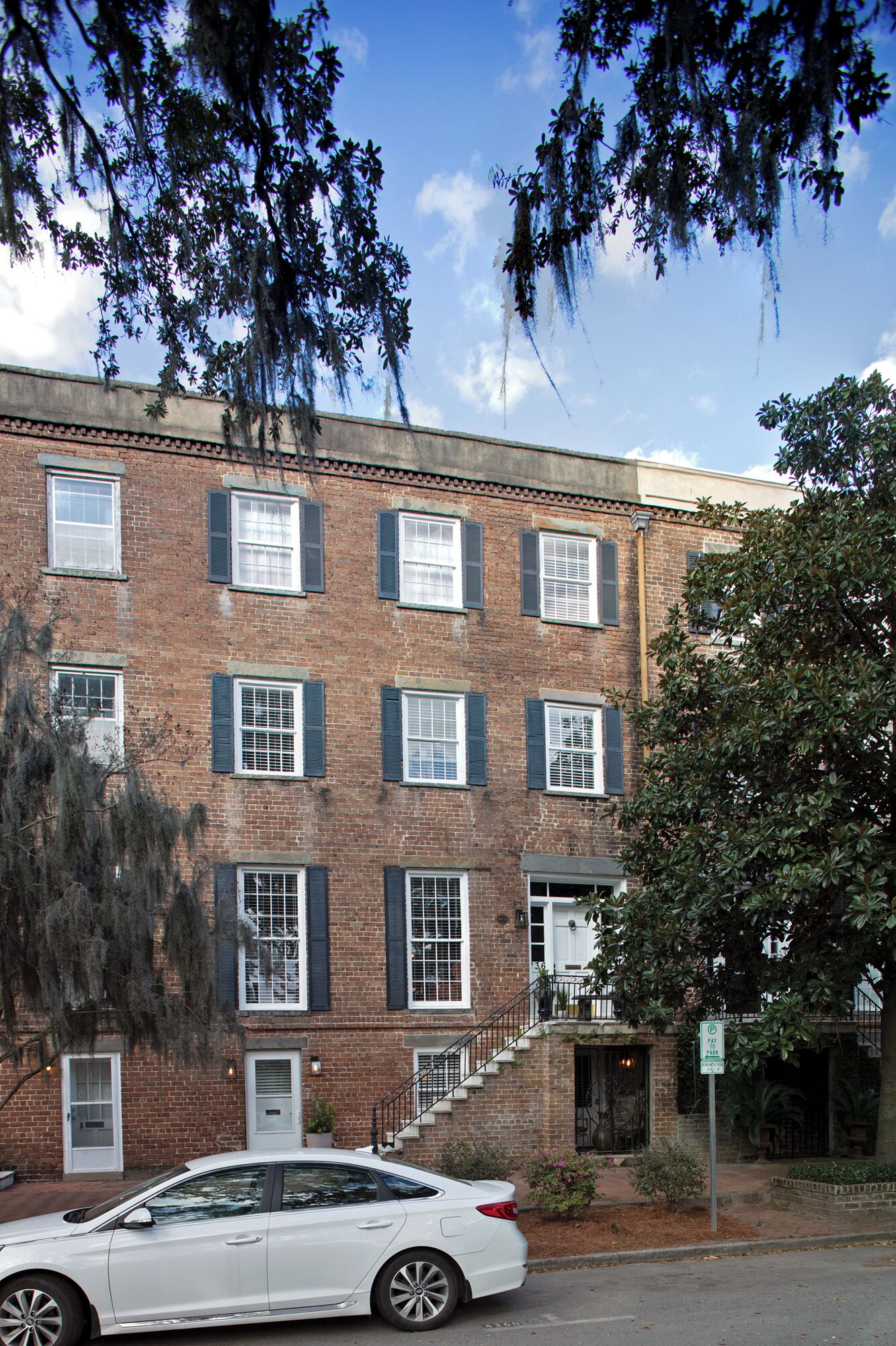 A two-story brick building with white-framed windows and shutters, a white car parked in front, and a tree with hanging moss.