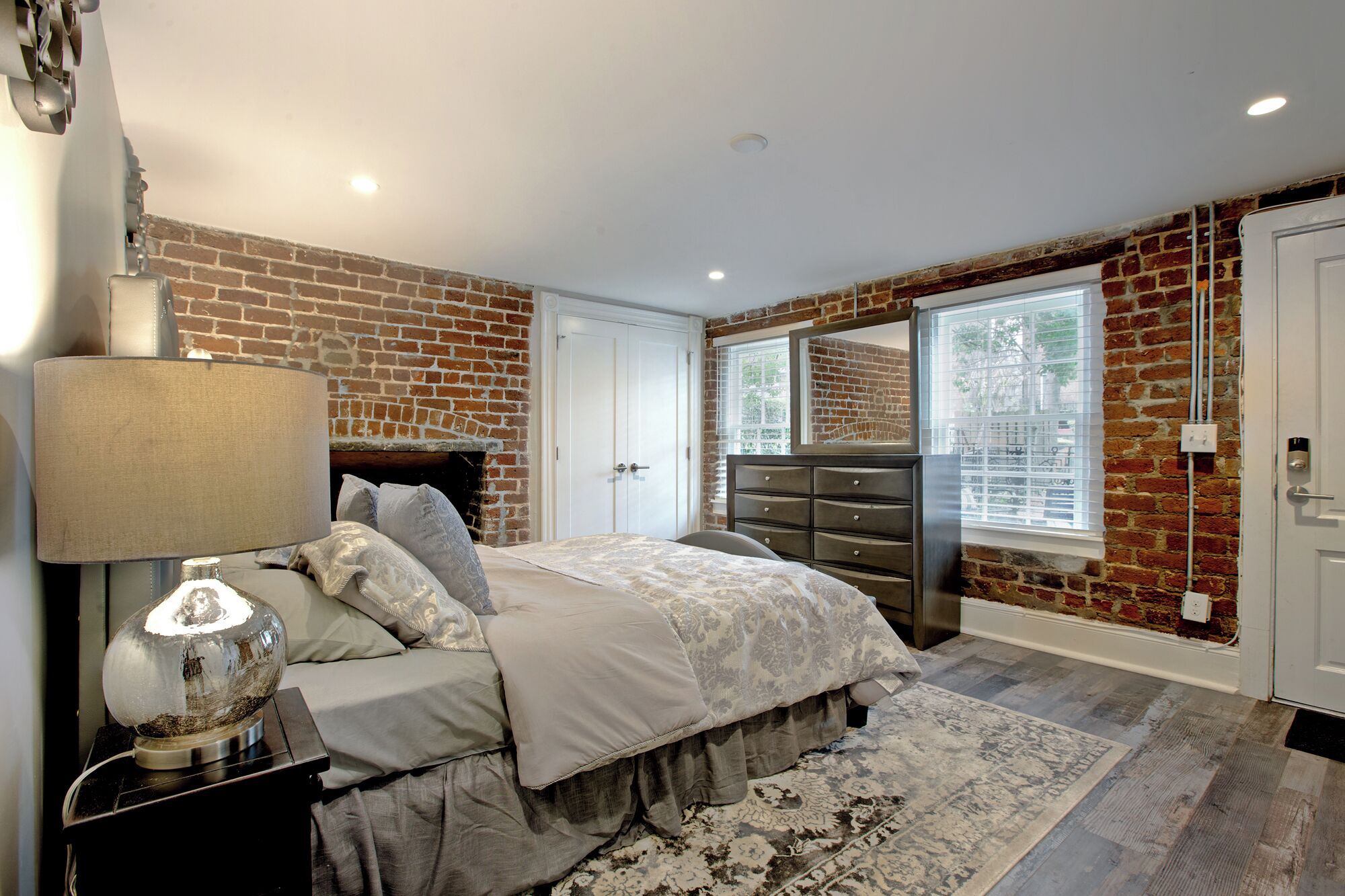 A bedroom with a brick wall, a bed with a grey duvet, a dresser, and a lamp.