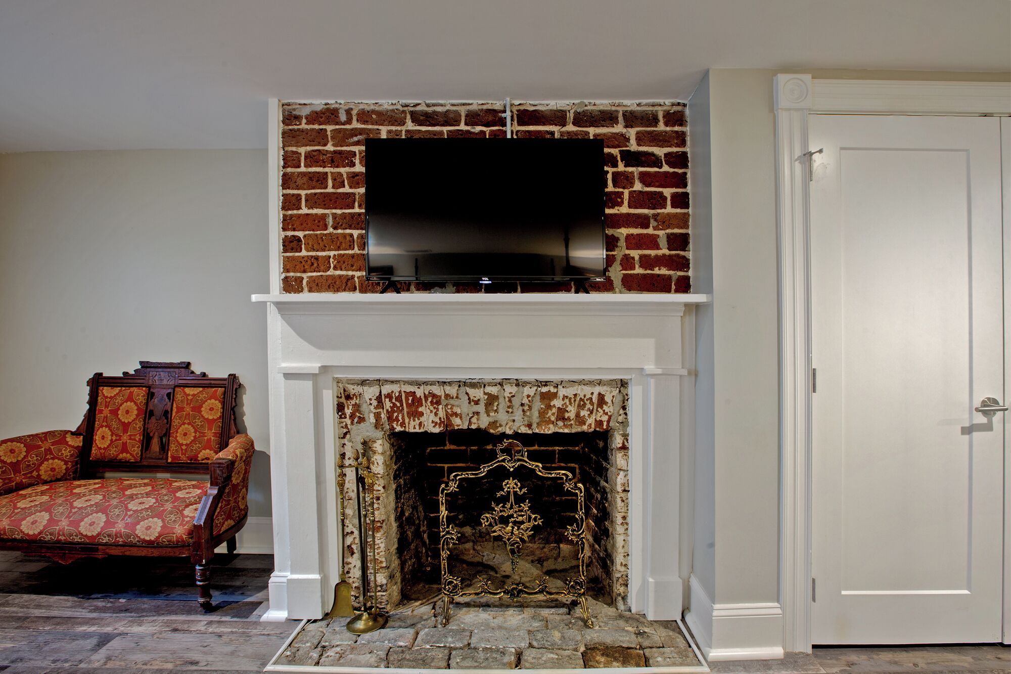 A room with a brick fireplace, a television mounted above it, a door to the right, and a patterned armchair to the left.