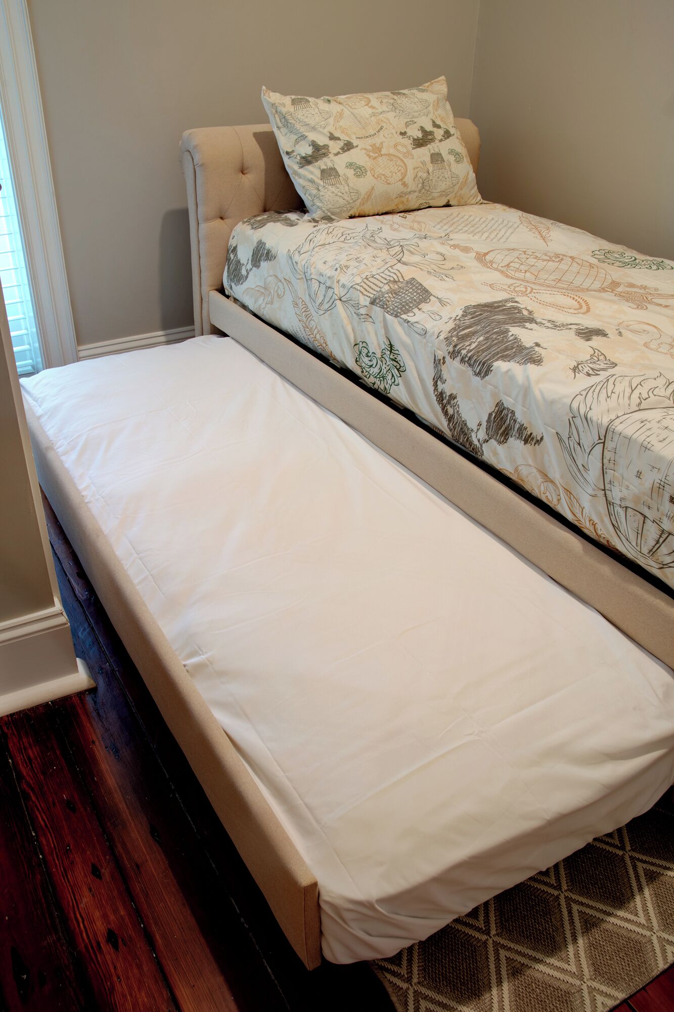 A single bed with a white mattress and a patterned duvet in a room with wooden flooring and a window with blinds.