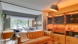 A modern bedroom with a large window, a sofa, a bedside table, and a built-in wooden cabinet with glass doors. - No. 1 Rosetree Suites - 3 Beds - Perfect Family Location in Calgary (Calgary)