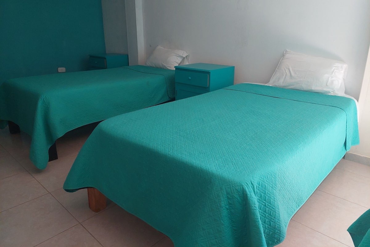 Ein Zimmer mit zwei Betten, beide mit türkisblau gestreifter Bettdecke und weißen Kissen. Zwei Nachttische, einer mit türkisblauer Schublade und der andere mit weißer Schublade. Die Wände sind weiß und der Boden ist mit Fliesen belegt.
