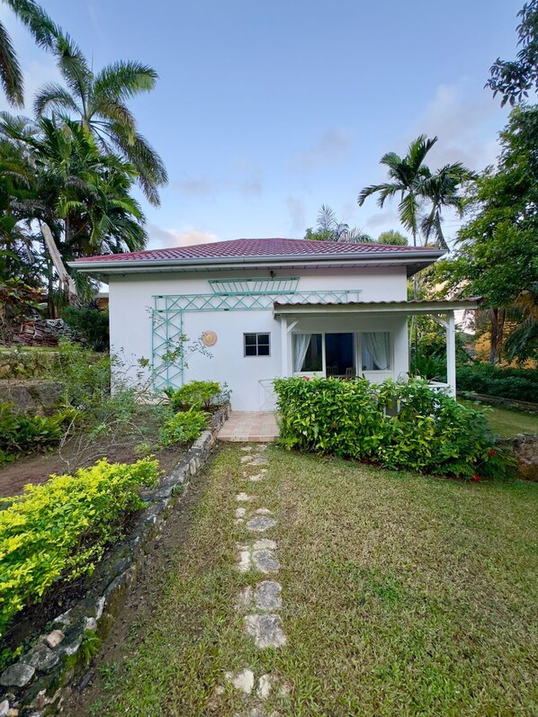 A small, single-story house with a tiled roof, surrounded by lush greenery and palm trees.