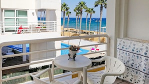 A balcony with white chairs and a small table, overlooking a pool and palm trees, with a view of the ocean.