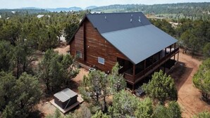 A wooden cabin with a large deck, surrounded by dense forest and mountains in the background.