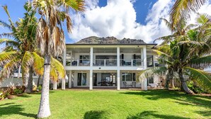 A two-story building with a covered patio area, surrounded by palm trees and a mountain in the background. - Tropical Oceanfront Paradise (Waianae)
