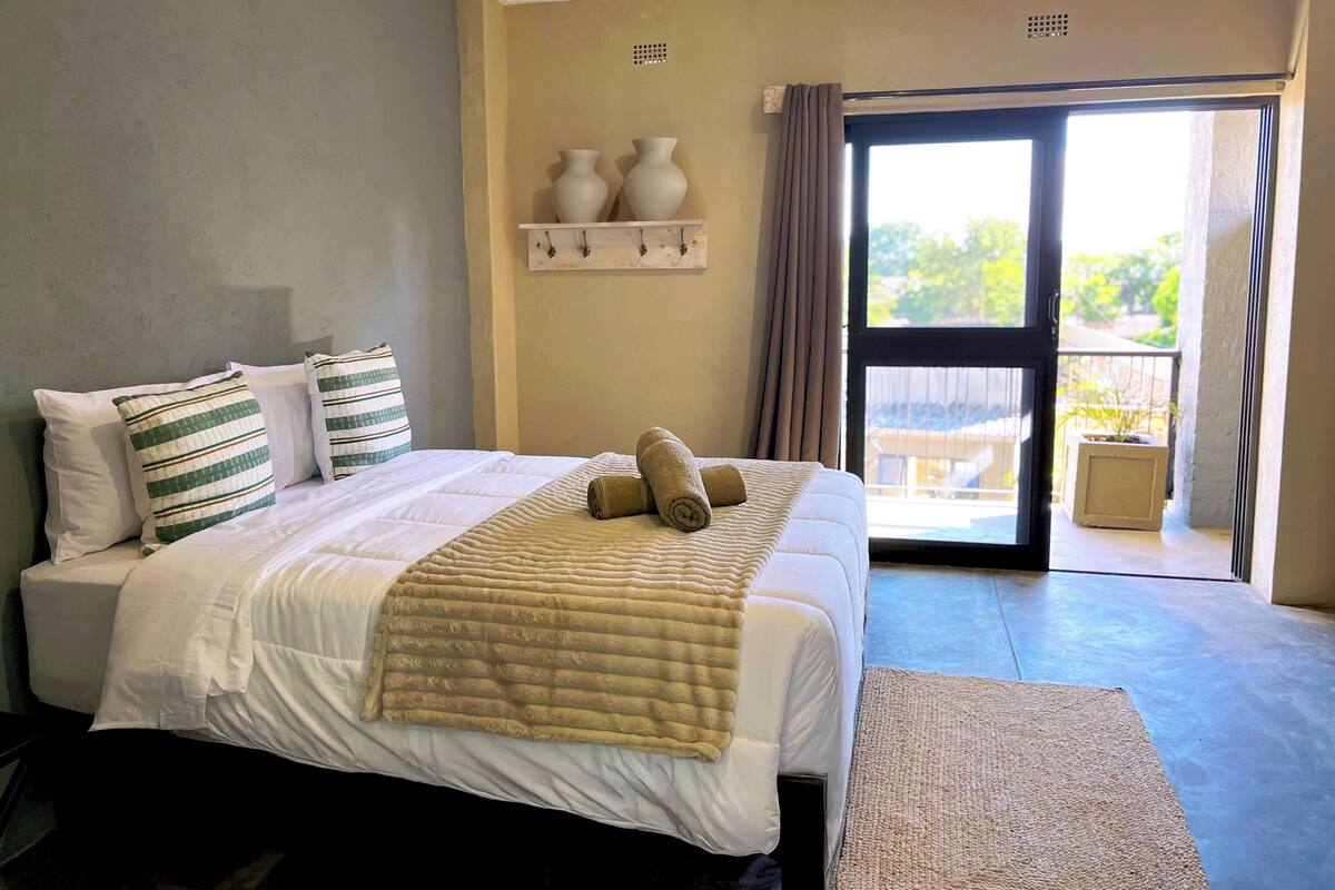 A bedroom with a bed, pillows, a bedside table, a window with curtains, and a view of the outdoors.