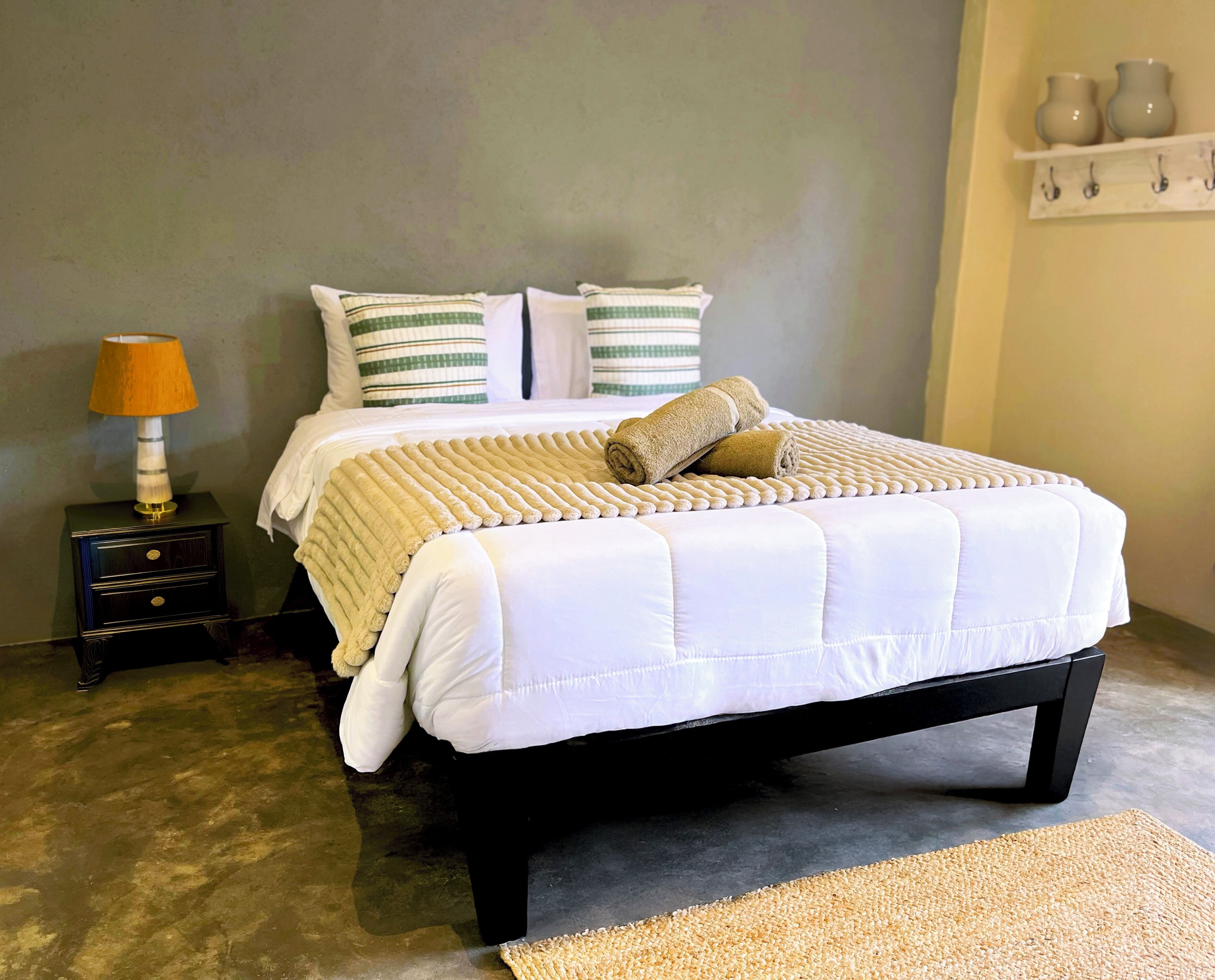 A neatly made bed with a white comforter and pillows, a small bedside table with a lamp, and a wall-mounted shelf with vases.