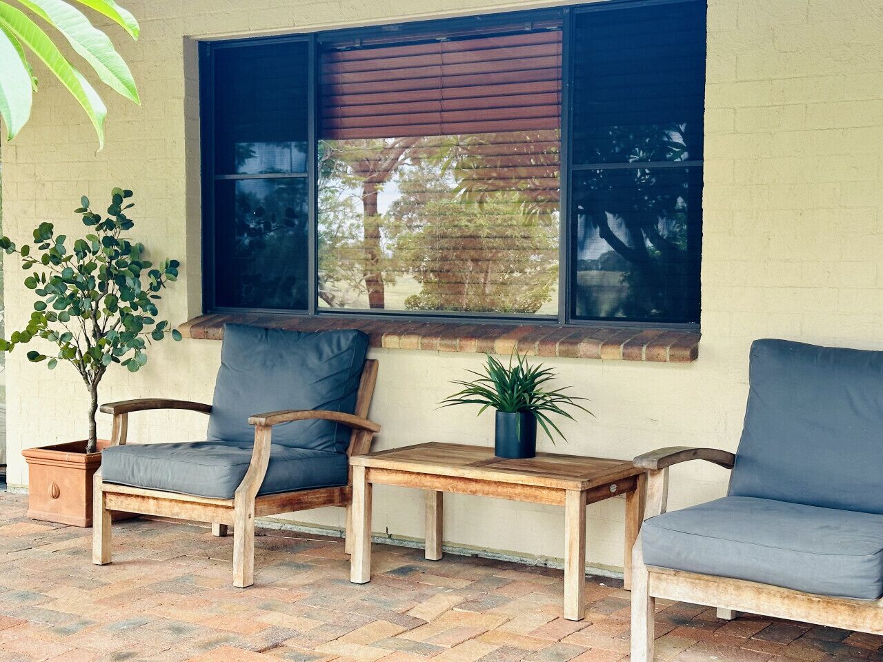 Une terrasse avec deux chaises en bois et une petite table, une plante en pot et une fenêtre avec des stores.