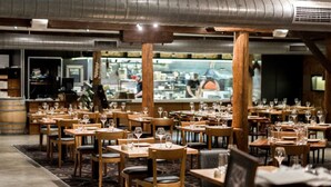 Een restaurantinterieur met meerdere tafels en stoelen, een bar op de achtergrond en een kamerbreed tapijt met patroon.